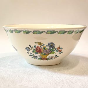 May include: A cream-colored ceramic bowl featuring a central floral basket motif with red, blue, purple, and yellow flowers. The rim is decorated with a continuous green leaf vine pattern. The bowl has a smooth, rounded shape and a wide opening.