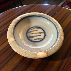 May include: A round ceramic bowl with a wide, textured rim. The interior features a smooth, off-white glaze with a dark blue circular band surrounding a central design of four horizontal brown brushstrokes. The exterior rim has a speckled finish.