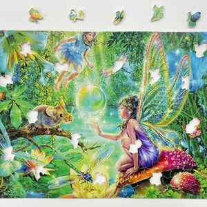 Pode incluir: Um quebra-cabeça colorido que apresenta uma fada com asas iridescentes sentada em um cogumelo, um rato, uma libélula e uma bolha. O quebra-cabeça é ambientado em uma floresta verde exuberante com flores e folhas.