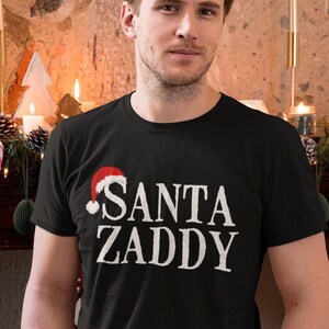 May include: A black t-shirt with a white graphic that says "SANTA ZADDY" with a red Santa hat on top of the "S".