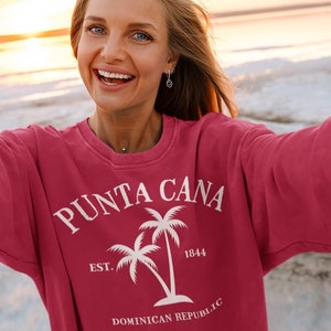 May include: A woman wearing a red sweatshirt with a white graphic design that says "Punta Cana Est. 1844 Dominican Republic" with two palm trees.