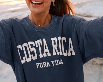 Peut inclure: Une femme portant un sweat-shirt bleu marine avec un texte blanc qui dit "Costa Rica Pura Vida" sur le devant. Elle sourit et prend un selfie sur une plage au coucher du soleil.