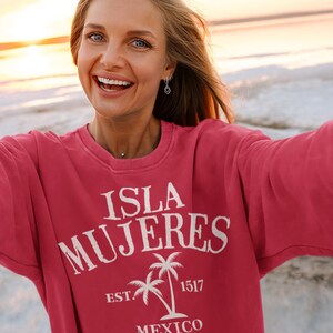 May include: A woman wearing a pink sweatshirt with a white graphic design that says "Isla Mujeres Est. 1517 Mexico" with a palm tree graphic. The woman is smiling and taking a selfie on a beach at sunset.