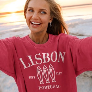 May include: A woman wearing a red sweatshirt with a white graphic design that says "LISBON EST. 1147 PORTUGAL" with three fish silhouettes. The woman is smiling and taking a selfie on a beach.