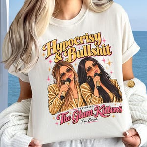 May include: A cream-colored t-shirt featuring a graphic of two women in gold sequin jackets holding microphones. The text reads Hypocrisy & Bullshit, The Glam Kittens, and I'm Bored, accented with pink and yellow starbursts.