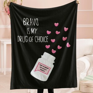 May include: Black fleece blanket with the text "BRAVO IS MY DRUG OF CHOICE" and a white pill bottle with the text "TAKE AS NEEDED FOR REALITY THERAPY". Pink hearts are scattered around the bottle.
