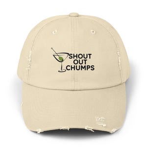 May include: A distressed, khaki-colored baseball cap with a curved brim. The cap features the text "SHOUT OUT CHUMPS" in black, along with a martini glass graphic with an olive. The cap has a worn, vintage look.