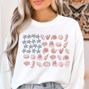 May include: White long-sleeve shirt with a red, white, and blue seashell and starfish design. The design resembles an American flag.