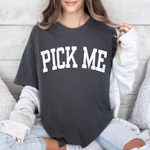May include: A dark gray t-shirt with the text "PICK ME" in white lettering. The shirt is worn by a person sitting on a couch.