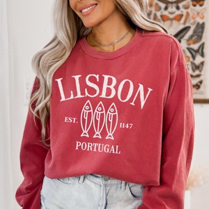May include: A red long-sleeved sweatshirt with a white graphic design that says "LISBON EST. 1147 PORTUGAL" with three stylized fish illustrations.