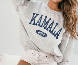 May include: A light gray sweatshirt with the word "KAMALA" in navy blue, arched across the chest. Below the name is the number "2024" in a navy blue oval. The sweatshirt is made of a soft material.