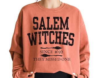 May include: A coral-coloured sweatshirt featuring the text "SALEM WITCHES" in bold black lettering. Below, it reads "SINCE 1692 THEY MISSED ONE" with broom illustrations. The sweatshirt has a crew neck and long sleeves.