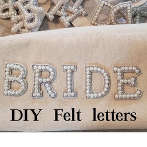 May include: DIY felt letters that spell out the word "BRIDE" with white pearl embellishments. The letters are on a beige canvas background.