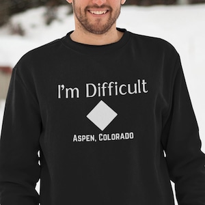 Könnte beinhalten: Schwarzes Sweatshirt mit dem Text "I'm Difficult" über einem weißen Diamanten und "ASPEN, COLORADO" darunter. Das Sweatshirt wird von einer Person in einer verschneiten Umgebung getragen.