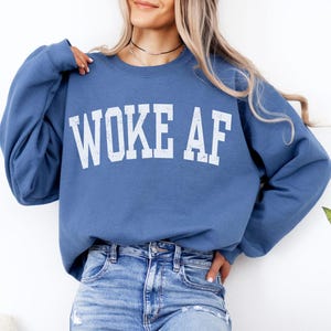 Könnte beinhalten: Ein blauer Pullover mit dem Aufdruck "WOKE AF" in Weiß auf der Vorderseite.