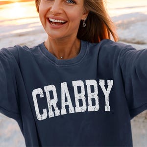 May include: Navy blue sweatshirt with the word "CRABBY" in large, distressed white letters. The sweatshirt has a crew neck and long sleeves. The fabric appears to be a soft, washed material.