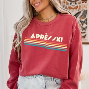 May include: A red long-sleeved sweatshirt with a retro-style graphic that says "APR&Egrave;S SKI" in white letters with a rainbow stripe design.