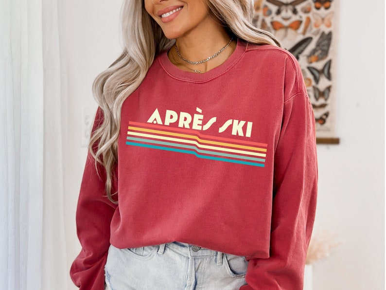May include: A red long-sleeved sweatshirt with a retro-style graphic that says "APR&Egrave;S SKI" in white letters with a rainbow stripe design.