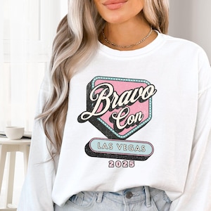 May include: White sweatshirt featuring a retro-style graphic for Bravo Con Las Vegas 2025. The design includes a pink and teal sign with the words "Bravo Con" in a vintage font, and "Las Vegas 2025" below.