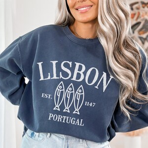 May include: A navy blue sweatshirt with a white graphic design that says "LISBON EST. 1147 PORTUGAL" with three stylized fish below the text.