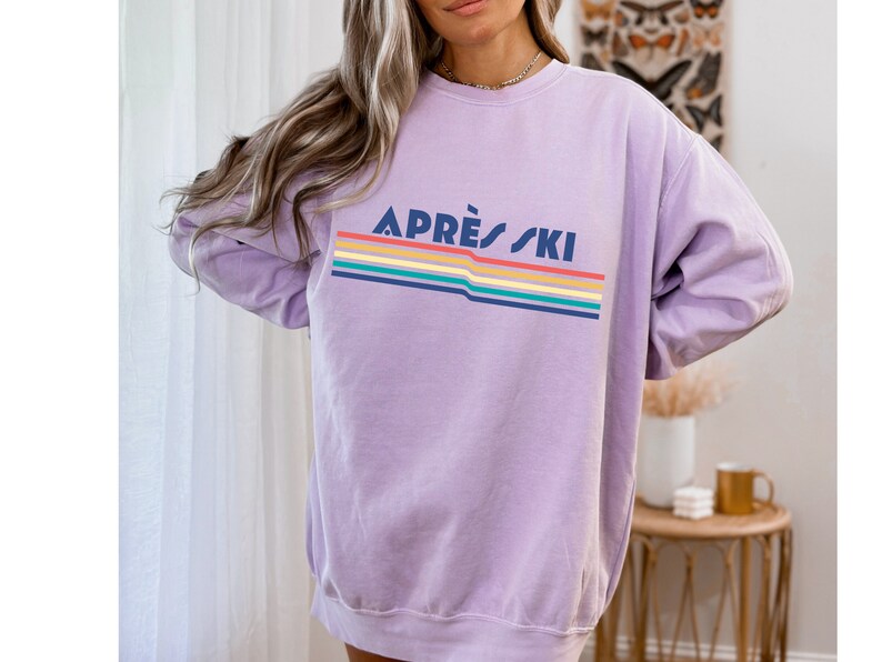 May include: A lavender sweatshirt with a rainbow stripe design and the text "APR&Egrave;S SKI" in navy blue.