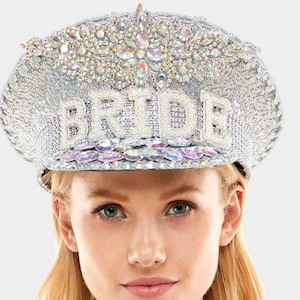 May include: A silver sequined hat with the word "BRIDE" spelled out in white beads. The hat is decorated with iridescent rhinestones and a large rhinestone flower.