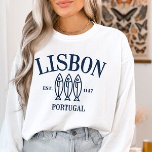 May include: White long-sleeve sweatshirt with a navy blue graphic print that says "LISBON EST. 1147 PORTUGAL" with three stylized fish.