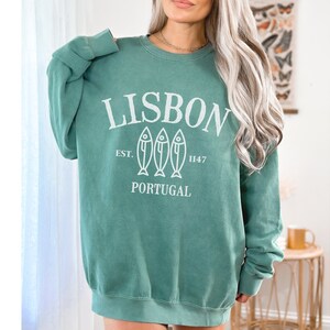 May include: A teal green sweatshirt with a white graphic print that says "LISBON EST. 1147 PORTUGAL" with three stylized fish below the text.