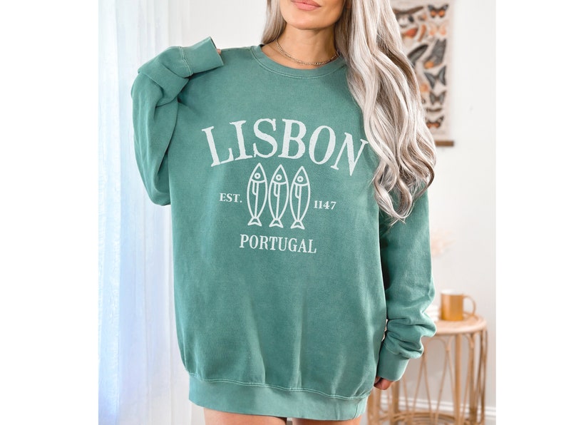 May include: A teal green sweatshirt with a white graphic print that says "LISBON EST. 1147 PORTUGAL" with three stylized fish below the text.