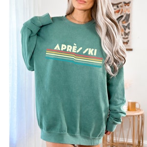 May include: A green sweatshirt with a retro-style rainbow stripe design and the text "APR&Egrave;S-SKI" in white.