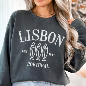 May include: A dark gray sweatshirt with a white graphic design that says "LISBON" with three fish illustrations and the text "EST. 1147 PORTUGAL".