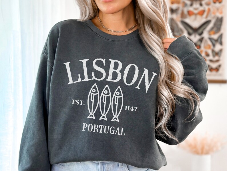 May include: A dark gray sweatshirt with a white graphic design that says "LISBON" with three fish illustrations and the text "EST. 1147 PORTUGAL".