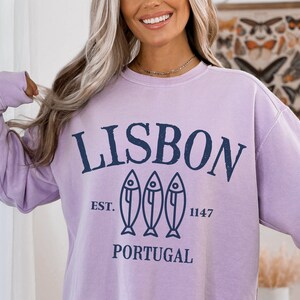 May include: A lavender sweatshirt with a navy blue graphic that says "LISBON" with three fish silhouettes and the text "EST. 1147 PORTUGAL".
