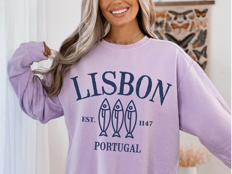 May include: A lavender sweatshirt with a navy blue graphic that says "LISBON" with three fish silhouettes and the text "EST. 1147 PORTUGAL".