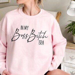 K&ouml;nnte beinhalten: Rosafarbener Pullover mit dem Aufdruck "In My Boss Bitch Era" in Schwarz.