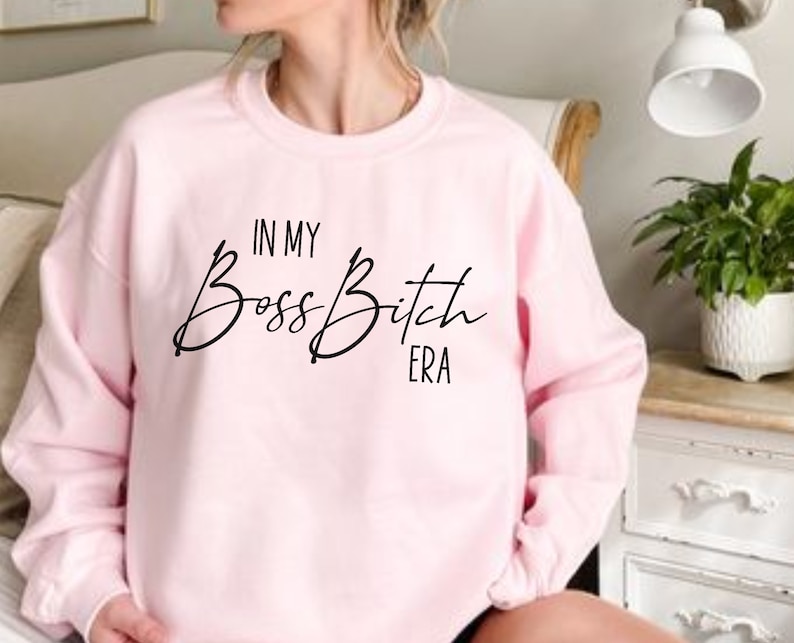 K&ouml;nnte beinhalten: Rosafarbener Pullover mit dem Aufdruck "In My Boss Bitch Era" in Schwarz.