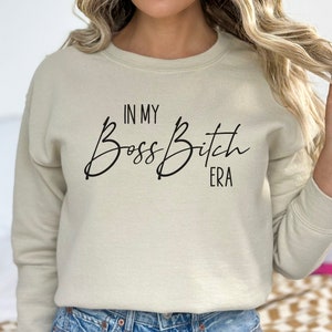 K&ouml;nnte beinhalten: Ein hellbeiger Pullover mit dem Aufdruck "In My Boss Bitch Era" in Schwarz.