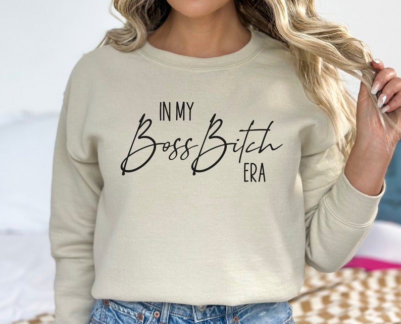K&ouml;nnte beinhalten: Ein hellbeiger Pullover mit dem Aufdruck "In My Boss Bitch Era" in Schwarz.