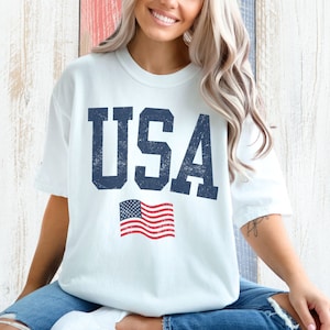 May include: A white t-shirt with a distressed blue "USA" graphic and a small American flag.