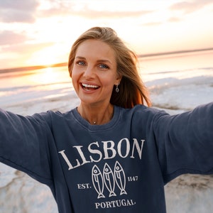 May include: A woman wearing a navy blue sweatshirt with a white graphic design that says "LISBON EST. 1147 PORTUGAL" with three fish silhouettes. The woman is smiling and taking a selfie on a beach at sunset.