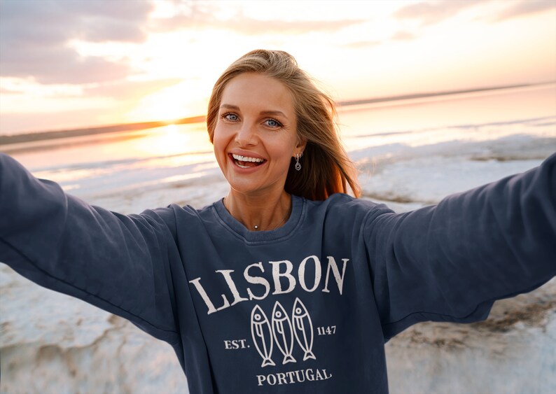 May include: A woman wearing a navy blue sweatshirt with a white graphic design that says "LISBON EST. 1147 PORTUGAL" with three fish silhouettes. The woman is smiling and taking a selfie on a beach at sunset.