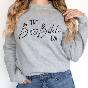 K&ouml;nnte beinhalten: Grauer Pullover mit dem Text "In My Boss Bitch Era" in schwarzer Kursivschrift.
