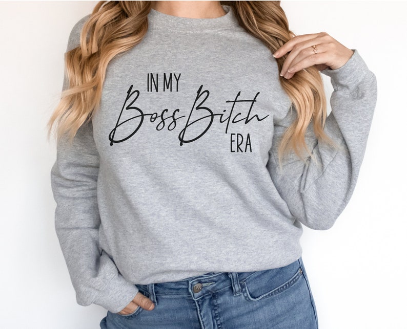 K&ouml;nnte beinhalten: Grauer Pullover mit dem Text "In My Boss Bitch Era" in schwarzer Kursivschrift.