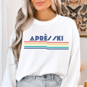 May include: White long-sleeve sweatshirt with a retro rainbow stripe design and the text "APR&Egrave;S SKI" in navy blue.