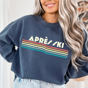 May include: A dark blue sweatshirt with a retro-style graphic that says "APR&Eacute;S SKI" in white letters with a rainbow stripe design.