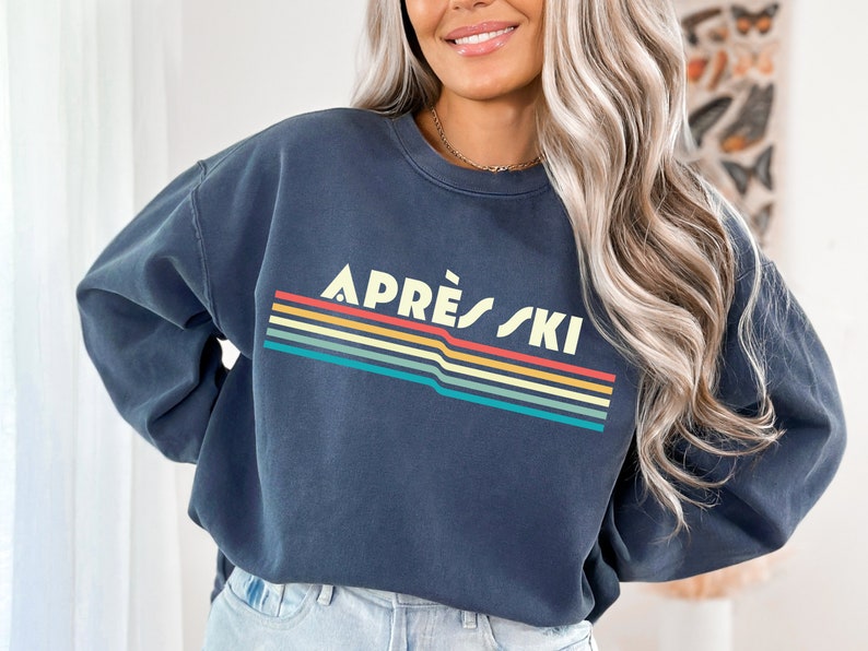 May include: A dark blue sweatshirt with a retro-style graphic that says "APR&Eacute;S SKI" in white letters with a rainbow stripe design.