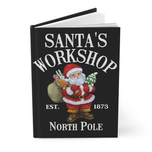 May include: A black journal with a white design featuring Santa Claus carrying a bag of toys and a Christmas tree. The text reads "Santa's Workshop Est. 1873 North Pole".