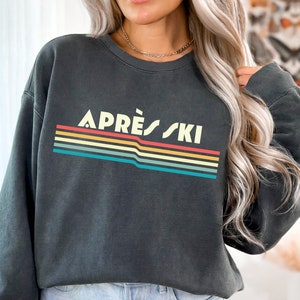 May include: A dark gray sweatshirt with a retro rainbow stripe design and the text "APR&Egrave;S SKI" in white.