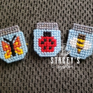 Può includere: Tre piccoli barattoli di tela plastica blu con sfondo bianco. Ogni barattolo ha un diverso disegno ricamato: una farfalla, una coccinella e un'ape. I barattoli sono su una superficie grigia testurizzata.