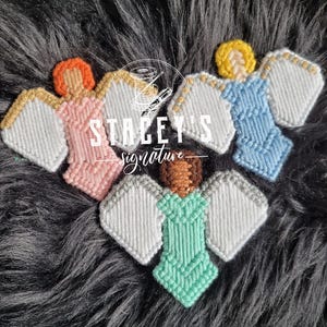 May include: Three handmade angel ornaments in pink, blue, and green, with white wings and various hair colours. The angels are made with a woven technique. The text "STACEY'S signature" is in the centre.